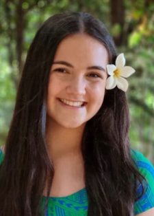A young woman with long dark hair smiles warmly. She has a white flower in her hair. The backdrop is a sunlit forest.