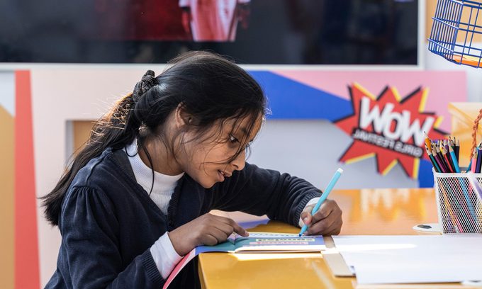 A girl concentrates on colouring at a bright table. A TV in the background shows a figure in white clothing