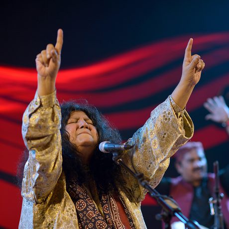 A musician with long hair passionately sings into a microphone, eyes closed, arms raised