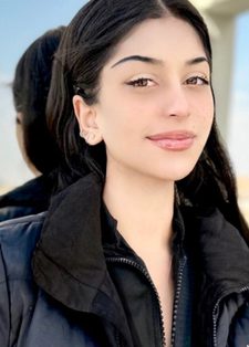 A young woman with long dark hair smiles slightly. She stands outside against a window on a sunny day.