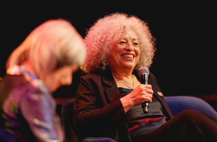 Angela Davis smiling and holding a microphone on stage. Another person in the foreground is slightly blurred. Warm, engaging atmosphere.
