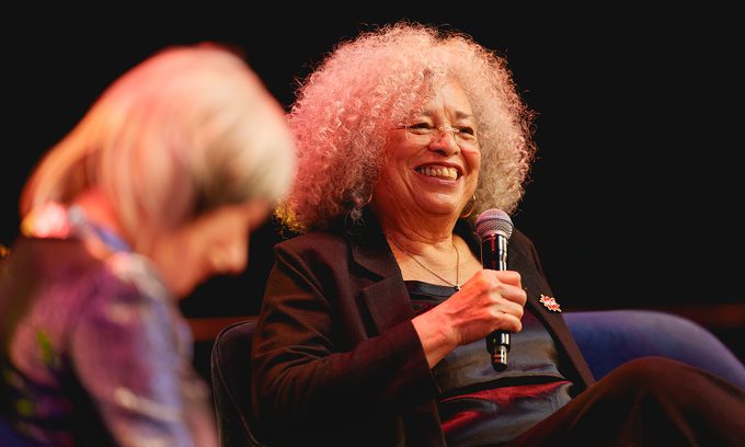 Angela Davis smiling and holding a microphone on stage. Another person in the foreground is slightly blurred. Warm, engaging atmosphere.