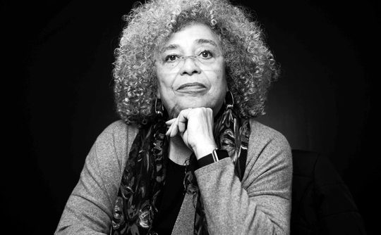 A black and white headshot of Angela Davis, a woman with grey afro hair and glasses, she is sat down with her hand on her chin.