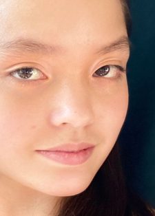 A young woman with long dark hair and subtle makeup looks calmly at the camera
