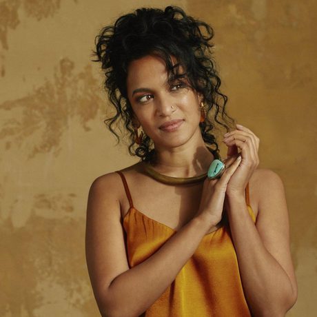 Anoushka Shankar looks to one side with her hands resting together. She has dark, curly hair tied up, and is wearing a shiny orange top and a ring with a large blue stone.