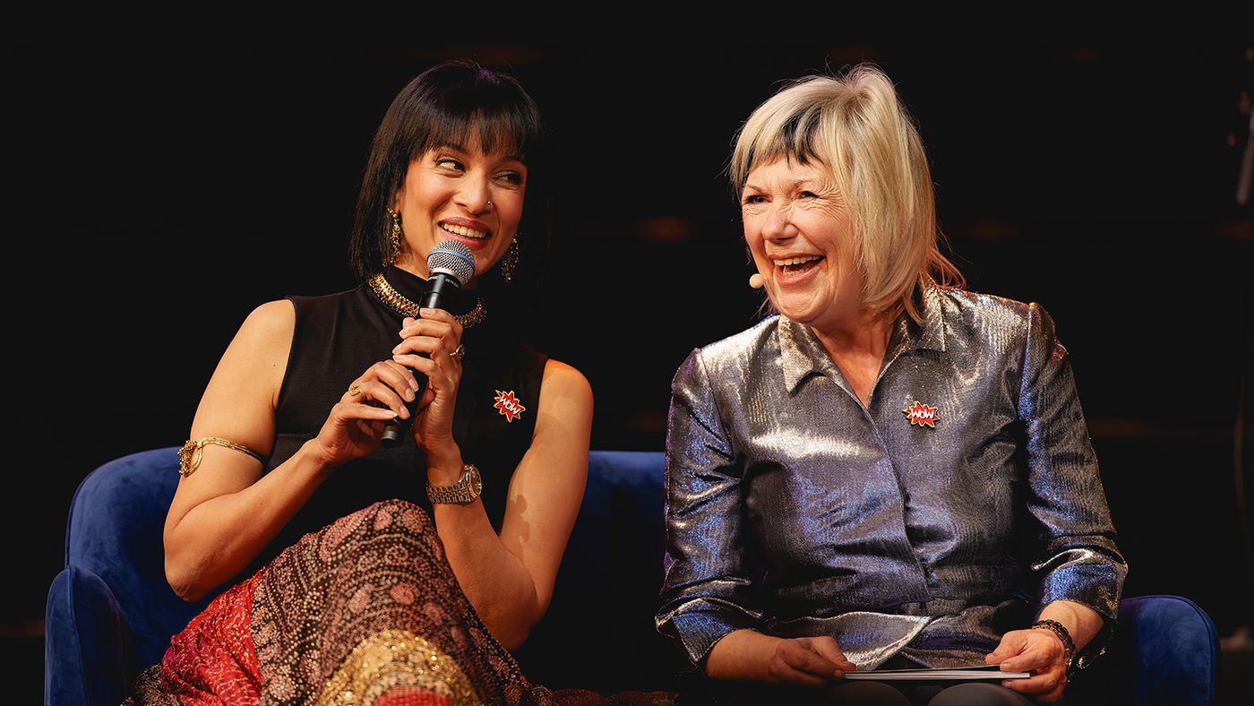 Two women sit on a blue couch, smiling warmly. The woman on the left holds a microphone and wears a patterned dress, while the woman on the right is in a shiny blouse. Both appear engaged and joyful.