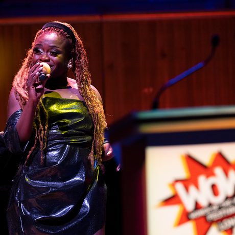 A singer with long braided hair performs on stage, holding a microphone. A podium with a "WOW" logo is visible in the foreground.