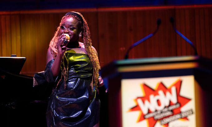 A singer with long braided hair performs on stage, holding a microphone. A podium with a "WOW" logo is visible in the foreground.