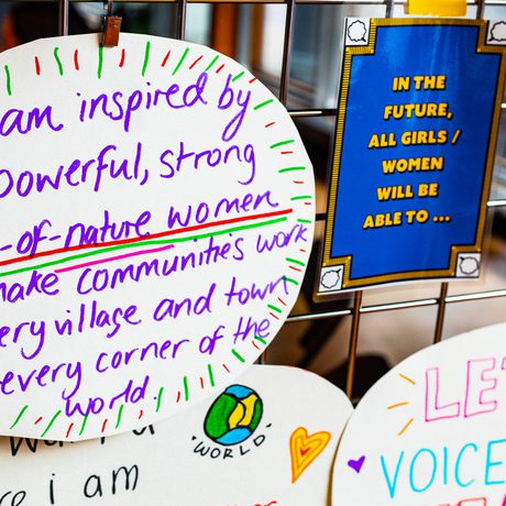 Decorated paper signs with empowering messages about women are clipped to a wire grid. A central sign reads, "I am inspired by the powerful, strong force-of-nature women who make communities work in every village and town in every corner of the world.""