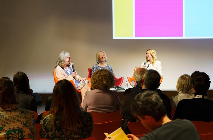 Three women seated on stage in a panel discussion, smiling and speaking into microphones. Audience members listen attentively. There is a bright, colourful background.