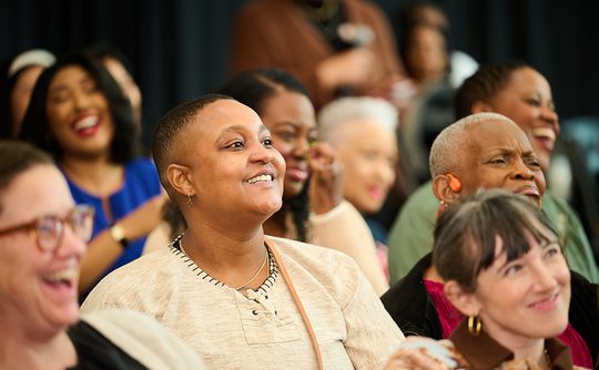 A diverse group of people seated indoors, smiling and laughing. The mood is joyful and lively