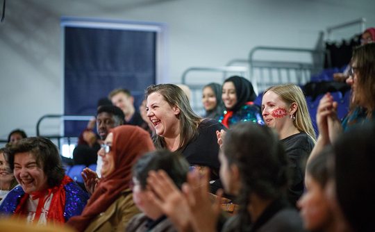 A diverse group of people are seated indoors, laughing and clapping. The atmosphere is joyful and lively