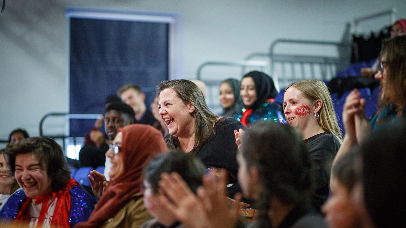 A diverse group of people are seated indoors, laughing and clapping. The atmosphere is joyful and lively