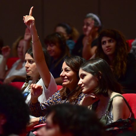 A diverse audience sits in a theatre with red seats, engaged in a presentation. A smiling woman raises her hand