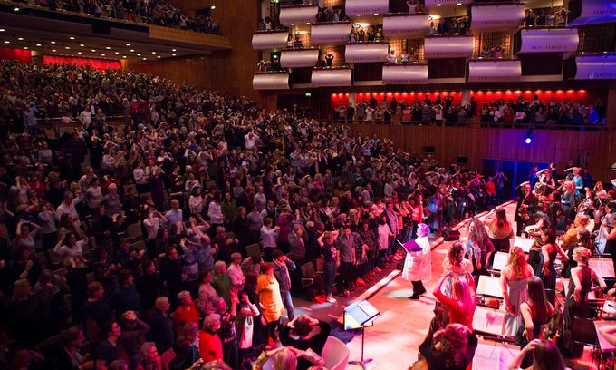 A vibrant concert hall is filled with a standing audience, applauding enthusiastically. The stage is lit warmly, showcasing musicians and performers.