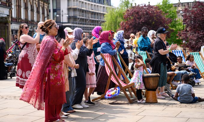 A diverse crowd claps and cheers at an outdoor event with deck chairs and a drum nearby. The scene is lively and joyful, set in a sunny urban area.