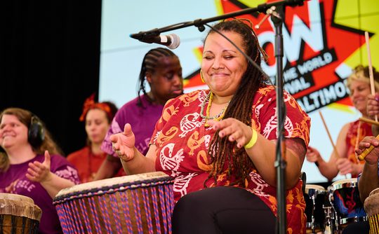 A group of diverse women joyfully play drums on stage. The central woman smiles with eyes closed. A banner in the background reads "WOW."