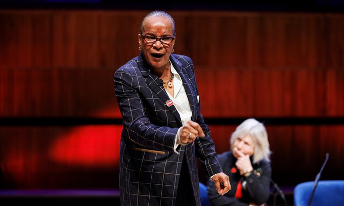 A person in a checkered suit speaks passionately on stage, gesturing with emphasis. Another person sits thoughtfully in the background