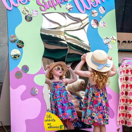 Young girl in a floral dress and sun hat stands before a colourful funhouse mirror. The Mirror reads "Women Supporting Women"