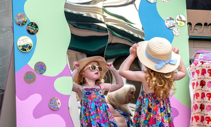 Young girl in a floral dress and sun hat stands before a colourful funhouse mirror. The Mirror reads "Women Supporting Women"