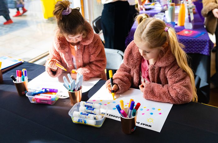 Two young girls in pink jackets focus on drawing with colourful markers at a table. Art supplies are scattered around.