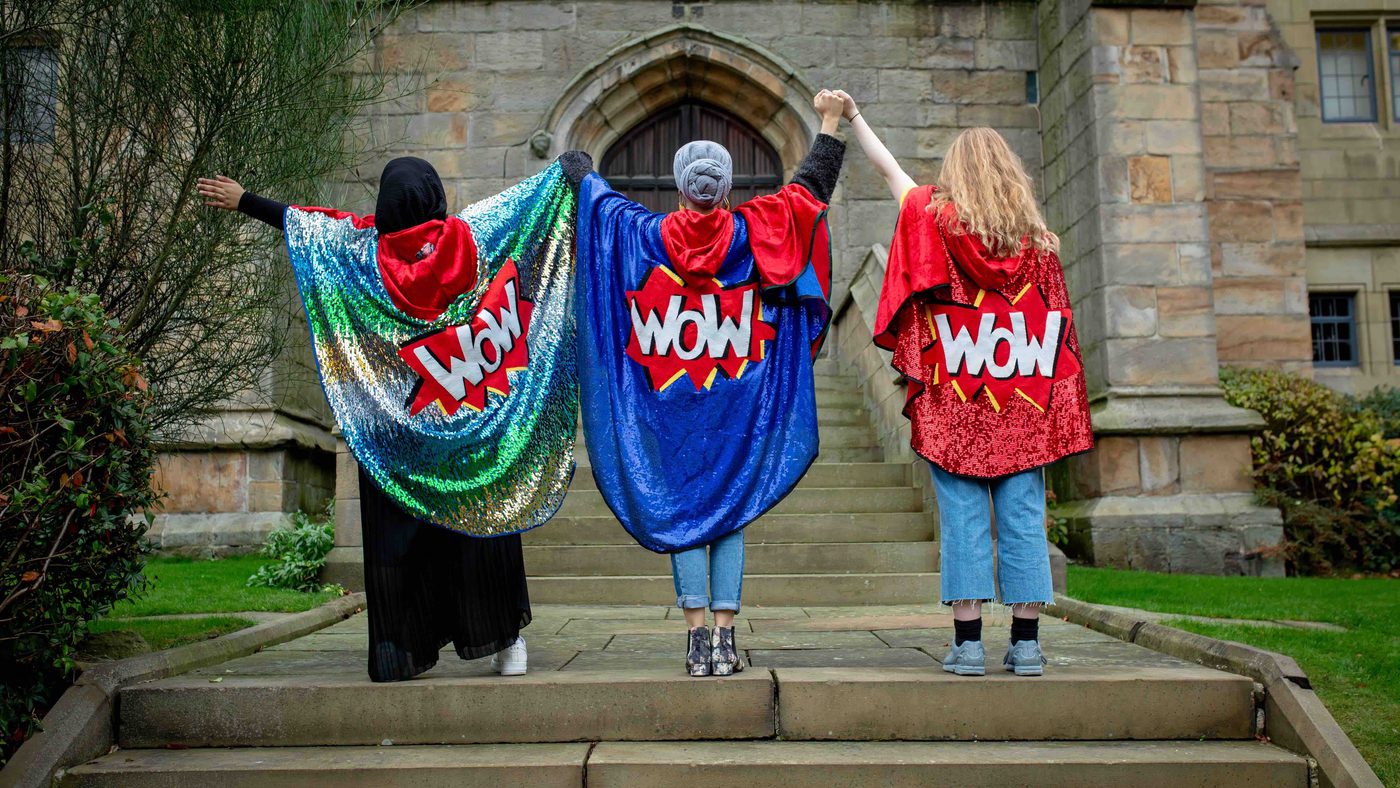 Three people stand on stone steps holding hands in the air, wearing colourful capes featuring the word "WOW." The mood is uplifting and empowering.