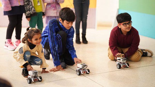 Three children, two boys and a girl, crouch on the floor with their fingers pressed on small robots with wheels.