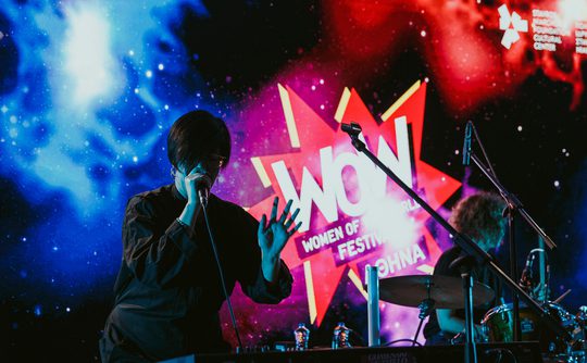 A performer sings passionately on stage at the "WOW Women of the World Festival." There is a colourful cosmic-themed backdrop