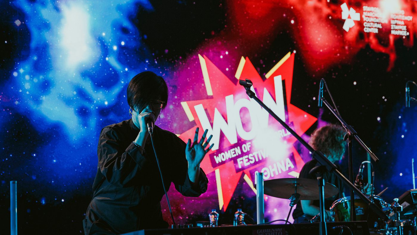 A performer sings passionately on stage at the "WOW Women of the World Festival." There is a colourful cosmic-themed backdrop