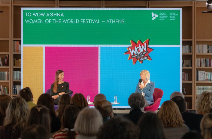 Two women engage in a discussion on stage at the WOW Festival in Athens. An audience listens attentively, with a vibrant backdrop displaying event details.