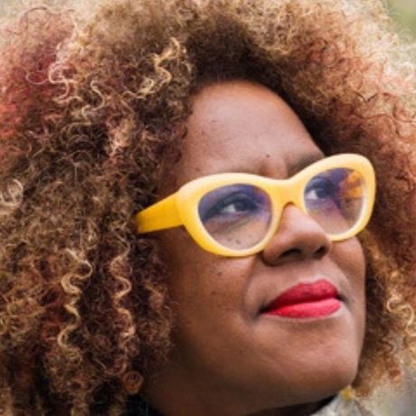 Headshot of Errollyn Wallen, who is looking off-camera. She has brown, red and blonde curly hair, and is wearing yellow glasses and red lipstick.