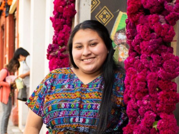 A woman in a colourful traditional dress smiles while standing next to vibrant magenta flowers on a lively street