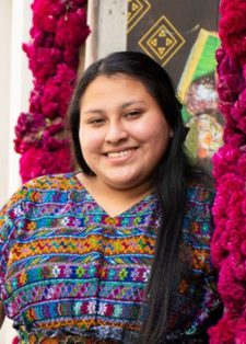 A woman in a colourful traditional dress smiles while standing next to vibrant magenta flowers on a lively street