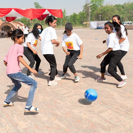 A group of six women play soccer with a blue ball on a sunny day. They smile, wearing casual clothes, with red and white banners in the background.