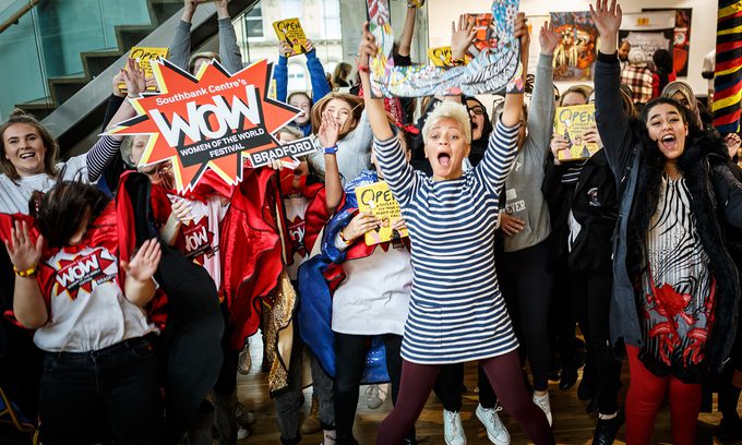 A diverse group of people celebrating energetically indoors. They hold colourful signs expressing joy and excitement.