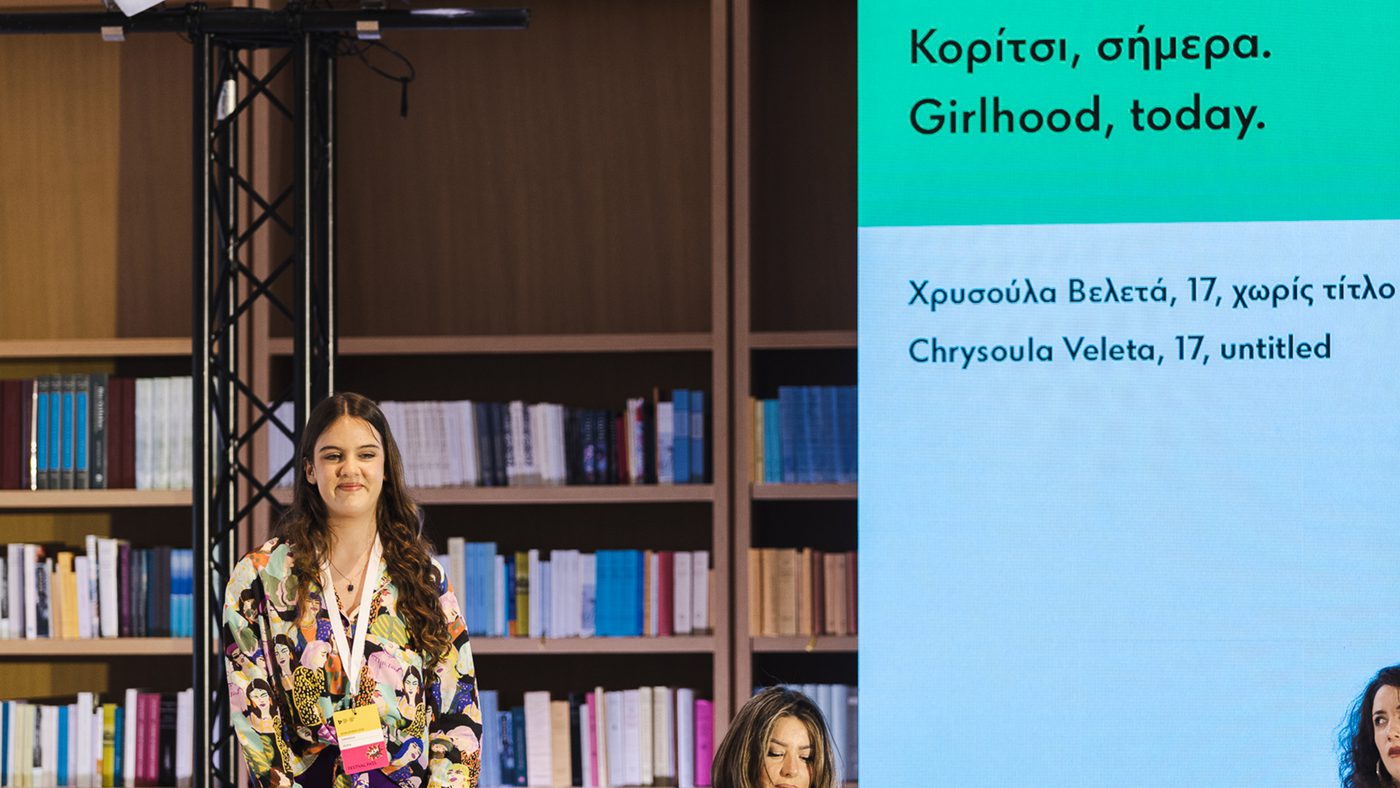 A young woman stands confidently on stage with a backdrop of bookshelves. Next to her, a large screen displays text about "Girlhood, today" in Greek and English.