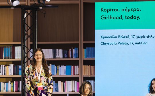 A young woman stands confidently on stage with a backdrop of bookshelves. Next to her, a large screen displays text about "Girlhood, today" in Greek and English.