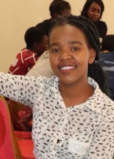 Young girl smiling, seated in a classroom setting. They wear a patterned white shirt. Behind, a group of students appear engaged in discussion.