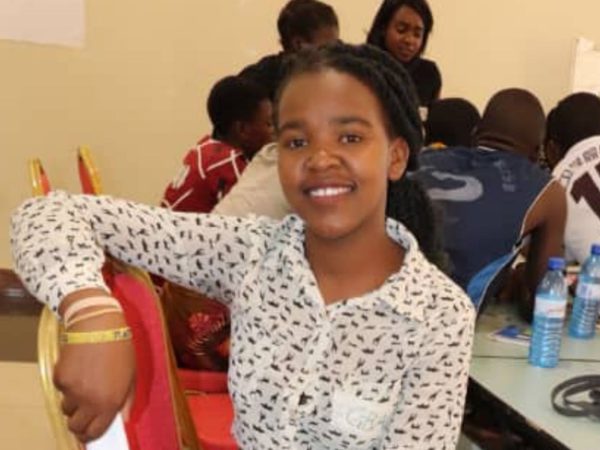 Young girl smiling, seated in a classroom setting. They wear a patterned white shirt. Behind, a group of students appear engaged in discussion.