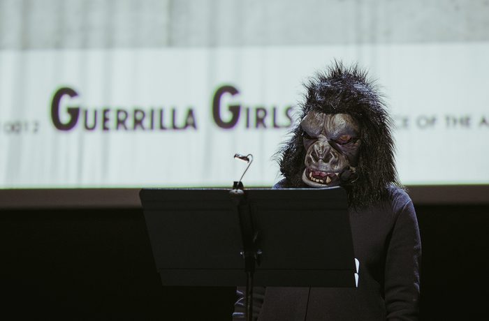 A person wearing a gorilla mask stands at a podium with a serious tone, in front of a screen displaying the words "Guerrilla Girls."