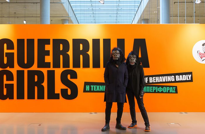 Two people wearing gorilla masks pose in front of large sign which says 'Guerrilla Girls'.
