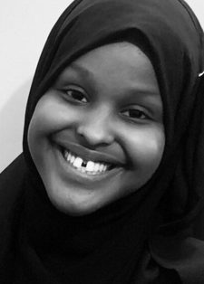 Black and white image of a young woman smiling wearing a dark hijab against a light background.