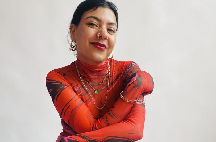A woman smiles at the camera with her arms crossed. She is wearing a red and black long-sleeved top and gold jewellery.
