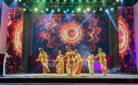 A group of performers dance on stage, framed by vibrant, intricate mandala patterns. The scene is lively and colourful, with bright stage lights.