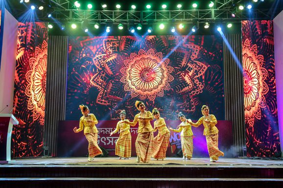 A group of performers dance on stage, framed by vibrant, intricate mandala patterns. The scene is lively and colourful, with bright stage lights.