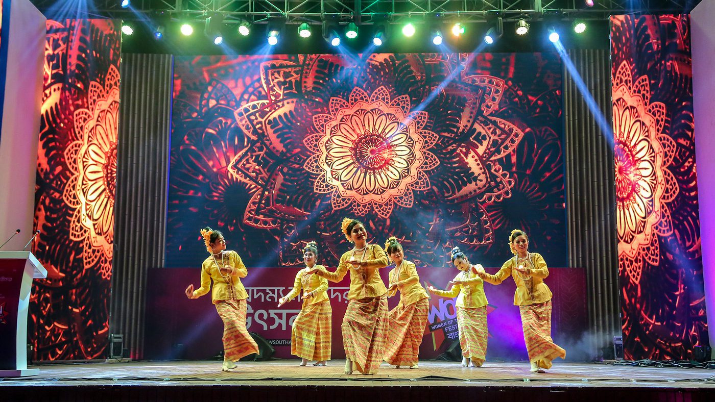 A group of performers dance on stage, framed by vibrant, intricate mandala patterns. The scene is lively and colourful, with bright stage lights.