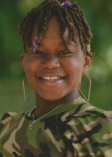 Smiling individual with short braided hair, wearing a camo-patterned shirt and hoop earrings, stands outdoors with blurred green foliage in the background.