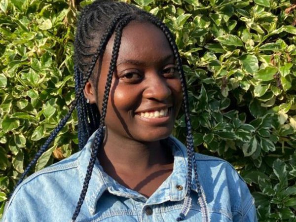 Smiling person with braided hair wearing a denim jacket stands in front of lush, green foliage