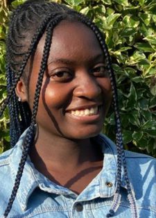 Smiling person with braided hair wearing a denim jacket stands in front of lush, green foliage