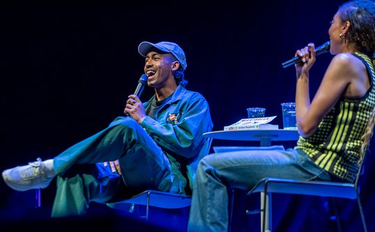 Two people sit on stage under blue lighting, holding microphones. Singer, Jordan Stephens is on the left, in a cap and blue outfit and appears to be laughing. The atmosphere is lively.